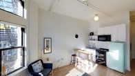 This single-floor space makes a striking impression with its dramatically vaulted ceilings and exposed ceiling beams, creating an airy, loft-like atmosphere that amplifies the sense of openness throughout. The interior is finished in a clean, neutral white palette that maximizes the natural light streaming in, lending the space a bright and welcoming ambiance.

The open-plan layout integrates the living and kitchen areas fluidly. The living zone features a comfortable armchair with decorative patterned cushions, accompanied by a petite side table and a sleek arc floor lamp — a curated, minimalist arrangement that makes efficient use of the space. A framed artwork adds a tasteful accent to the white wall.

The kitchen is compact yet well-equipped, featuring open shelving, a gas range, a microwave oven, and a sink with ample counter space. A small wooden dining table paired with stools sits adjacent to the kitchen, offering a casual dining area that bridges both zones seamlessly.

Warm hardwood flooring runs throughout, adding texture and warmth to the otherwise crisp, minimal aesthetic. The combination of soaring ceilings, white walls, natural wood tones, and thoughtfully selected furnishings gives the space a modern, Scandinavian-inspired character that is both functional and visually refined.