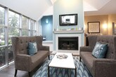 This single-floor space is designed with an open-concept layout that combines a comfortable lounge area, a dining bar, and a well-appointed wet bar station into one cohesive environment. The soaring vaulted ceilings lend an airy, expansive feel throughout, while rich hardwood flooring provides warmth and continuity across the space.

The lounge area is anchored by a classic fireplace with a full mantle and a wall-mounted flat-screen television above, set against a soft teal accent wall that adds a touch of color. A tufted wingback loveseat, accent chair, and a low coffee table create an inviting seating arrangement centered on a patterned area rug.

Floor-to-ceiling windows wrap around one side of the space, flooding the interior with abundant natural light and offering sweeping views of lush, tree-lined outdoor surroundings. A long counter-height bar with sleek stools runs directly along these windows, making it an ideal spot to enjoy the scenery.

A dedicated wet bar area features dark cabinetry, a built-in beverage cooler, and an eye-catching tiled accent wall framed by wall sconces and tasteful artwork — adding both functionality and style.

French doors with frosted glass panes provide access to an adjacent room, maintaining an open feel while offering flexibility for privacy. The overall ambiance is refined yet welcoming, blending modern amenities with comfortable, residential warmth.