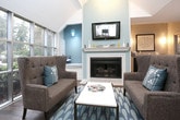 This single-floor space is designed with an open-concept layout that combines a comfortable lounge area, a dining bar, and a well-appointed wet bar station into one cohesive environment. The soaring vaulted ceilings lend an airy, expansive feel throughout, while rich hardwood flooring provides warmth and continuity across the space.

The lounge area is anchored by a classic fireplace with a full mantle and a wall-mounted flat-screen television above, set against a soft teal accent wall that adds a touch of color. A tufted wingback loveseat, accent chair, and a low coffee table create an inviting seating arrangement centered on a patterned area rug.

Floor-to-ceiling windows wrap around one side of the space, flooding the interior with abundant natural light and offering sweeping views of lush, tree-lined outdoor surroundings. A long counter-height bar with sleek stools runs directly along these windows, making it an ideal spot to enjoy the scenery.

A dedicated wet bar area features dark cabinetry, a built-in beverage cooler, and an eye-catching tiled accent wall framed by wall sconces and tasteful artwork — adding both functionality and style.

French doors with frosted glass panes provide access to an adjacent room, maintaining an open feel while offering flexibility for privacy. The overall ambiance is refined yet welcoming, blending modern amenities with comfortable, residential warmth.