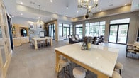 This single-story clubhouse is designed with a contemporary aesthetic, offering a welcoming and versatile environment for socializing and recreation. The main gathering area is expansive and open-concept, anchored by large marble-topped tables with natural wood bases and surrounded by upholstered bar stools, making it ideal for group activities or casual meetings. Elegant chandelier lighting adds a refined touch overhead, while recessed lighting ensures the space remains bright throughout.

A comfortable lounge area features dark upholstered seating arranged around a central rug, creating an inviting setting for relaxed conversation. Floor-to-ceiling black-framed windows and glass doors line the perimeter, flooding the interior with natural light and offering views of the outdoor surroundings. Large-format tile flooring in a neutral gray tone runs consistently throughout, lending the space a cohesive, polished look.

Adjacent to the main area, a well-appointed refreshment bar features light wood cabinetry, quartz countertops, a mosaic tile backsplash, and a commercial coffee station. A glass-walled fitness center is also accessible from the main floor, housing a variety of exercise equipment visible through its transparent walls.