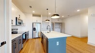 This single-floor unit showcases a clean, modern design throughout, with light-toned hardwood flooring and crisp white walls creating a bright, cohesive aesthetic. The open-concept kitchen serves as the heart of the space, anchored by a substantial center island with a light blue base and white quartz countertop, an integrated sink, and stylish matte black pendant lighting overhead. Stainless steel appliances, including a French door refrigerator, range, and dishwasher, line the perimeter alongside tall white cabinetry and a subway tile backsplash. A dedicated laundry area is conveniently accessible just off the kitchen.

The bedrooms feature plush carpeting and large windows that invite generous natural light and outdoor views of the surrounding neighborhood. One room offers direct access to a balcony through a full-height glass door. A spacious walk-in closet with wire shelving and carpeted flooring provides practical storage. An additional room with hardwood flooring and a window offers flexibility for various uses.

The bathroom is finished with a white vanity, quartz countertop, matte black hardware and fixtures, a large framed mirror, and a glass-enclosed tiled shower, reinforcing the unit's consistent contemporary style.