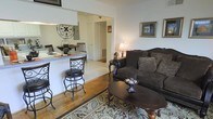 This single-floor home offers a thoughtfully designed layout that blends comfort and functionality. The living area serves as the heart of the space, featuring rich hardwood flooring, a plush dark sofa adorned with decorative pillows, and a classic oval coffee table set atop a patterned area rug. Warm ambient lighting from a traditional table lamp creates a cozy and welcoming atmosphere, complemented by framed artwork displayed on the walls.

Flowing directly from the living area is an open-concept kitchen and dining space, separated by a raised breakfast bar with elegant wrought iron bar stools. The kitchen counter provides a casual dining option, while a dining table with wooden chairs is visible beyond, making the space well-suited for both everyday meals and entertaining guests. Light tile flooring in the kitchen area contrasts beautifully with the hardwood of the living room, subtly defining each zone.

A decorative wrought iron wall piece adds a distinctive artistic touch to the kitchen wall, contributing to the home's classic and curated aesthetic. A hallway visible in the background suggests additional rooms beyond, further extending the functional footprint of this well-appointed single-story home.