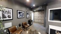 This single-floor space exudes a contemporary, urban character defined by a carefully curated mix of materials and finishes. The walls are painted in a deep charcoal gray, lending the interior a dramatic and intimate atmosphere that is complemented by recessed ceiling lighting and an industrial-style geometric pendant light. A striking horizontal wood-plank accent wall introduces warm, natural texture that contrasts effectively against the cooler gray tones throughout the space.

The seating area features cognac leather chairs paired with a sleek black side cabinet, creating a stylish yet functional arrangement. A cowhide rug anchors the seating zone, adding an organic, eclectic element to the design. Black-framed monochromatic portrait photographs hang prominently on the walls, reinforcing the space's bold, artistic identity. A small gold wire accent table adds a subtle metallic detail to the composition.

Carpeted flooring in a neutral, textured pattern runs throughout, providing both comfort and visual cohesion. The overall ambiance is polished and intentional, blending industrial and contemporary design elements to create a refined, moody environment well-suited for a professional or creative setting.