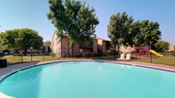 This outdoor amenity space centers around a generously sized swimming pool with striking turquoise water, creating a refreshing and inviting atmosphere. The pool area is finished with a clean, light-colored concrete deck that surrounds the water on multiple sides, providing ample space for relaxation. A pair of classic lounge chairs are positioned poolside, offering a comfortable spot to unwind and take in the surroundings.

The space is enclosed by a black metal fence, providing both security and a clean visual boundary that defines the pool area. Several mature, full-canopy trees frame the background, offering natural shade and a lush, green setting that enhances the overall ambiance. The landscaping contributes a sense of tranquility and privacy to the space.

In the background, multi-story brick residential buildings with wood-accented balconies and staircases are visible, constructed in a warm, earthy tone that complements the natural greenery. The architecture features a traditional style with pitched rooflines. The clear blue sky overhead reinforces the bright, open, and pleasant character of this outdoor communal amenity space.