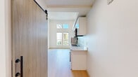 Located at 4612 Stone Way N in Seattle's vibrant Wallingford neighborhood, this efficiently designed studio unit makes a strong impression through its striking architectural details. The space features soaring double-height ceilings that create an open, loft-like atmosphere, enhanced by tall vertical windows and clerestory windows above that flood the interior with natural light and views of the surrounding greenery.

The kitchen is thoughtfully outfitted with sleek white cabinetry extending floor to ceiling, a stainless steel refrigerator, an integrated cooktop, stainless steel sink with a modern gooseneck faucet, built-in microwave, and a dishwasher. An in-unit washer is also conveniently included. Light-toned wood-style flooring runs throughout the main living area, complementing the clean, minimalist aesthetic of the white walls and contemporary finishes.

A sliding barn-style door provides access to the main living space, adding a stylish architectural accent. The bathroom features a clean, all-white walk-in shower with a built-in shelf niche and chrome fixtures. An upper mezzanine-level railing is visible from the main floor, contributing to the vertical drama of the space. A bold red entry door adds a pop of color and character to the otherwise crisp, modern interior palette.