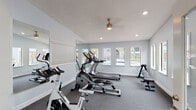 Located at 3132 W Lambright St in Leto, FL, this single-floor fitness center is designed to support a well-rounded cardio and strength training routine. The main workout area features gray carpeted flooring and is outfitted with a selection of equipment, including a treadmill, spinning bike, elliptical trainer, and a weight bench. Large wall-mounted mirrors line the perimeter, visually expanding the space and allowing users to monitor their form during exercise. Recessed lighting and a ceiling fan provide consistent illumination and airflow throughout the room. Multiple windows along the walls flood the interior with natural light and offer views of the outdoor pool area, creating an energizing atmosphere. The light gray walls and textured ceiling contribute to a clean, functional aesthetic. A tiled entryway leads into the main fitness area, providing a clear transition point upon entry. A security camera and posted fitness guidelines reflect a commitment to safety and proper facility use. The space is accessible via a keyed exterior door and is equipped with an emergency exit, ensuring compliance with safety standards. Overall, this fitness center offers a bright, well-equipped environment suited for daily exercise.