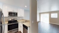 This 1-bedroom, 1-bathroom residence is designed for comfortable, efficient living across a single floor. The kitchen is outfitted with stainless steel appliances, including an electric range, built-in microwave, and dishwasher, complemented by crisp white shaker cabinetry and light-toned countertops. A stainless steel sink sits beneath recessed lighting, adding a clean, modern touch. A structural column subtly defines the transition between the kitchen and the adjacent living area, creating a sense of distinct zones while maintaining an open feel. The living space features smooth, neutral-toned walls and hard-surface flooring, lending the area a sleek and low-maintenance quality. A sliding glass door with vertical blinds floods the living area with natural light and offers a connection to the outdoors. The textured ceiling adds a layer of architectural detail throughout. Overall, the unit presents a clean, neutral palette that provides a versatile backdrop for a variety of interior styles, making it a practical and inviting space for everyday living.