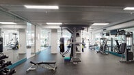Located at 3530 E La Palma Ave in Anaheim, California, this single-floor facility is designed to support both physical fitness and community wellness. The space features a well-appointed fitness center with rubber flooring, equipped with a multi-functional cable training tower, a heavy punching bag, adjustable weight benches, cardio machines, and cable equipment. Large floor-to-ceiling windows line the gym area, inviting abundant natural light and providing views of the outdoor surroundings, while wall-mounted mirrors enhance the sense of openness. Recessed and panel lighting throughout ensures a bright, functional environment. Adjacent to the fitness area, glass-walled rooms create transparent, inviting gathering spaces. One area serves as a casual lounge, furnished with plaid armchairs and wicker seating arranged around a light hardwood floor, bathed in natural light from expansive windows. Another glass-enclosed room functions as a library or activity space, featuring built-in shelving with books and colorful items, and casual seating. A clean, modern aesthetic is carried throughout the facility, with a consistent white and light gray color palette accented by teal trim details, polished columns, and terrazzo-style flooring in the common corridors.