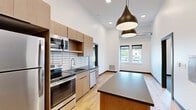Located at 2939 Main St in Buffalo, NY, this thoughtfully designed unit offers a modern aesthetic across a well-utilized single floor. The kitchen serves as a standout feature, showcasing warm wood cabinetry, dark countertops, a white subway tile backsplash, and a full suite of stainless steel appliances including a refrigerator, range, microwave, and dishwasher. A central island with a dark countertop provides additional workspace and adds to the kitchen's functional appeal. Dramatic pendant lighting hangs from notably tall ceilings, enhancing the open and airy ambiance of the space. Light-toned hardwood flooring flows through the kitchen and living areas, complementing the clean white walls throughout. The additional rooms are finished with neutral carpet and feature windows that bring in natural light and exterior views, keeping the spaces bright and inviting. The bathroom includes a wood vanity with dark countertop, a large mirror, and a tub-shower combination with clean, simple fixtures. A second vanity area with ample counter space and drawers adds a practical touch. The overall design balances contemporary finishes with functional layouts, making this a well-appointed space in the heart of Buffalo.