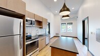 Located at 2939 Main St in Buffalo, NY, this thoughtfully designed unit offers a modern aesthetic across a well-utilized single floor. The kitchen serves as a standout feature, showcasing warm wood cabinetry, dark countertops, a white subway tile backsplash, and a full suite of stainless steel appliances including a refrigerator, range, microwave, and dishwasher. A central island with a dark countertop provides additional workspace and adds to the kitchen's functional appeal. Dramatic pendant lighting hangs from notably tall ceilings, enhancing the open and airy ambiance of the space. Light-toned hardwood flooring flows through the kitchen and living areas, complementing the clean white walls throughout. The additional rooms are finished with neutral carpet and feature windows that bring in natural light and exterior views, keeping the spaces bright and inviting. The bathroom includes a wood vanity with dark countertop, a large mirror, and a tub-shower combination with clean, simple fixtures. A second vanity area with ample counter space and drawers adds a practical touch. The overall design balances contemporary finishes with functional layouts, making this a well-appointed space in the heart of Buffalo.
