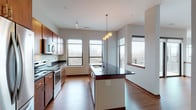 This well-appointed single-floor residence offers a thoughtfully designed open-concept layout that seamlessly connects the kitchen and living areas. The kitchen is a standout feature, showcasing rich dark granite countertops, warm-toned wood cabinetry, and a full suite of stainless steel appliances including a range, microwave, and dishwasher. A central island with an integrated sink adds both functionality and visual appeal, while a stylish pendant light fixture anchors the space above it. Hardwood flooring flows throughout the main living areas, complemented by dark wood baseboards that add a refined touch. Multiple large windows flood the space with natural light and offer elevated views of the surrounding landscape and neighborhood. A private balcony extends the living space outdoors. The two bedrooms are finished with plush carpeting and feature generous closet space, including a built-in shelved closet in one room. Both bathrooms are well-equipped — one features a sliding glass-door walk-in shower, while the other includes a tub-shower combination — each with tile flooring, wood-toned vanities, and stone-topped counters. Overall, the space presents a clean, contemporary aesthetic with warm wood accents throughout.
