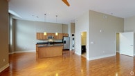 This well-designed single-floor space showcases a thoughtful open-concept layout that seamlessly connects the kitchen and living areas. The kitchen is a standout feature, outfitted with warm honey-toned wood cabinetry, dark granite countertops, and a full suite of stainless steel appliances, including a refrigerator with ice and water dispenser and a built-in microwave. A central kitchen island provides additional prep space and casual seating, while two pendant lights hang above, adding a modern touch. Tall ceilings amplify the sense of openness, and a ceiling fan keeps the space comfortable year-round. Rich hardwood floors run throughout, reflecting light beautifully and tying the space together. Neutral sage-gray wall tones create a calm, contemporary atmosphere. A dedicated in-unit laundry area with a stacked washer and dryer is conveniently tucked just off the main living space, adding everyday practicality. A doorway visible from the main area leads to what appears to be a bathroom, completing the functional layout. The overall ambiance is clean, bright, and modern — ideal for those seeking a comfortable and efficient living environment.