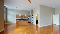 This well-designed single-floor space showcases a thoughtful open-concept layout that seamlessly connects the kitchen and living areas. The kitchen is a standout feature, outfitted with warm honey-toned wood cabinetry, dark granite countertops, and a full suite of stainless steel appliances, including a refrigerator with ice and water dispenser and a built-in microwave. A central kitchen island provides additional prep space and casual seating, while two pendant lights hang above, adding a modern touch. Tall ceilings amplify the sense of openness, and a ceiling fan keeps the space comfortable year-round. Rich hardwood floors run throughout, reflecting light beautifully and tying the space together. Neutral sage-gray wall tones create a calm, contemporary atmosphere. A dedicated in-unit laundry area with a stacked washer and dryer is conveniently tucked just off the main living space, adding everyday practicality. A doorway visible from the main area leads to what appears to be a bathroom, completing the functional layout. The overall ambiance is clean, bright, and modern — ideal for those seeking a comfortable and efficient living environment.