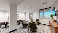 Located at 76 N Winchester Blvd in Santa Clara, CA, this single-floor commercial space combines contemporary design with mid-century modern sensibility to create a sophisticated and functional environment. The main lounge area is anchored by a distinctive plaid-patterned carpet that adds visual texture throughout the space, complemented by crisp white walls and recessed lighting that ensures a bright, welcoming atmosphere.

The seating area features a curated arrangement of green upholstered lounge chairs with walnut-toned frames, centered around a compact side table — ideal for informal meetings or relaxed conversation. A separate dining or small group workspace is furnished with a round table and coordinating upholstered chairs, offering additional flexibility for collaborative use.

Framed landscape artwork adorns the walls, lending a refined aesthetic, while large windows fitted with roller shades allow natural light to filter in and provide views of the surrounding greenery. A textured accent wall near the entrance adds depth and visual interest, decorated with circular wall art pieces.

A well-appointed built-in bookcase with dark shelving displays curated books and decorative objects, adding warmth and character. A warm-toned wooden credenza completes the space, offering practical storage. Together, these elements create a polished, inviting environment well-suited for professional or hospitality use.