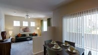 This single-floor residence offers a well-designed open-concept layout that seamlessly connects the living and dining areas, creating a cohesive and functional living environment. The living room is carpeted and comfortably appointed, featuring a ceiling fan with integrated lighting and multiple windows dressed with white blinds and green curtain panels that invite generous amounts of natural light. A low half-wall partition elegantly separates the living area from the dining space, maintaining an open and airy feel while providing a subtle sense of definition between the two zones. The dining area is positioned near a large sliding glass door fitted with vertical blinds, allowing additional light to flood the space and offering potential access to an outdoor area. Warm, neutral-toned walls throughout lend a calm and inviting ambiance, while the overall layout promotes easy flow and everyday practicality. The combination of soft carpeting in the living room, ample window coverage, and thoughtful spatial arrangement makes this a comfortable and welcoming single-level home suited for both relaxed everyday living and casual entertaining.