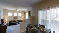 This single-floor residence offers a well-designed open-concept layout that seamlessly connects the living and dining areas, creating a cohesive and functional living environment. The living room is carpeted and comfortably appointed, featuring a ceiling fan with integrated lighting and multiple windows dressed with white blinds and green curtain panels that invite generous amounts of natural light. A low half-wall partition elegantly separates the living area from the dining space, maintaining an open and airy feel while providing a subtle sense of definition between the two zones. The dining area is positioned near a large sliding glass door fitted with vertical blinds, allowing additional light to flood the space and offering potential access to an outdoor area. Warm, neutral-toned walls throughout lend a calm and inviting ambiance, while the overall layout promotes easy flow and everyday practicality. The combination of soft carpeting in the living room, ample window coverage, and thoughtful spatial arrangement makes this a comfortable and welcoming single-level home suited for both relaxed everyday living and casual entertaining.