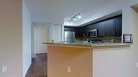 This single-floor space offers a thoughtfully designed interior that blends modern aesthetics with functional living. The kitchen serves as a central highlight, featuring rich dark cabinetry that contrasts elegantly with granite countertops. A raised breakfast bar provides a casual dining or workspace option, while the mosaic tile backsplash adds a decorative layer of texture and depth to the cooking area. Stainless steel appliances, including a full-size refrigerator and built-in microwave, bring a sleek, contemporary finish to the space. Track lighting overhead ensures the kitchen is well-lit and practical. The flooring throughout is a warm hardwood-style material that adds continuity and warmth across the living areas. A hallway visible from the kitchen leads to additional rooms, suggesting a well-organized layout that balances open communal areas with private spaces. The clean white walls and neutral color palette create a bright, inviting atmosphere throughout. Overall, the space presents a modern and cohesive design that is both visually appealing and highly functional for everyday living.