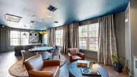 Located in Orlando, FL, this thoughtfully designed single-floor lounge space makes a striking first impression with its bold, deep teal ceiling that anchors the entire interior with a sense of drama and sophistication. The living room serves as the heart of the space, combining a media area and a casual seating zone seamlessly. Built-in cabinetry in a soft sage-green finish supports a large wall-mounted display, while rich cognac leather armchairs and a round dark wood coffee table create an inviting conversation area. A circular dining arrangement with upholstered seating and an ornate area rug adds a refined social element. The herringbone and geometric-patterned hardwood-style flooring throughout lends visual texture and cohesion. Large multi-pane windows dressed with elegant linen drapes flood the space with natural light and offer views of lush, tropical greenery outside. A wet bar corner, adorned with botanical wallpaper and fitted with a sink and under-counter storage, adds a functional and decorative touch. Curated wall art displayed in panel-molded frames enhances the gallery-like atmosphere. The connecting hallway transitions smoothly from the living area, maintaining the same refined palette and design continuity throughout this well-appointed space.