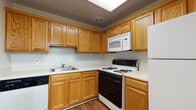 This single-floor residence offers a practical and comfortable layout spread across several well-defined living areas. The kitchen serves as a functional hub, featuring warm oak cabinetry extending from floor to ceiling, providing generous storage throughout. White appliances include a gas range with a built-in overhead microwave, a dishwasher, and a refrigerator, all complemented by light countertops and a stainless steel sink. The flooring transitions between hardwood-style and carpet depending on the area, adding visual contrast throughout the unit.

The living area is notably open and bright, with soft carpeting underfoot and a large sliding glass door fitted with vertical blinds that allow abundant natural light to fill the space. The entry area features a hardwood-style floor transition at the front door, creating a defined entryway.

The bedrooms are carpeted and well-proportioned, each equipped with ceiling fans and lighting fixtures. One bedroom features access to closet storage as well as an adjoining room, potentially a walk-in closet or en suite. Windows with blinds allow natural light into the sleeping areas while maintaining privacy. Overall, the space presents a clean, neutral palette suitable for a variety of furnishing styles.
