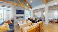 Located at 9001 Blue Flag Dr in Simpsonville, SC, this single-story community clubhouse blends coastal-inspired design with warm, inviting details throughout. The open floor plan is anchored by a dramatic lounge area showcasing soaring vaulted ceilings with exposed natural wood beams painted in a soft blue, complemented by a bold patterned tile feature wall housing a linear fireplace and wall-mounted television. Large glass doors flood the space with natural light and provide direct access to an outdoor patio. The kitchen area is a standout, featuring a generous marble-look island with seating, sage-toned lower cabinetry, a striking blue Moroccan-pattern tile backsplash, open shelving, and oversized brass pendant lights that add a glamorous finishing touch. A full-size French door refrigerator and built-in appliances complete the functional yet stylish space. A casual dining and lounge nook is defined by exposed brick accent walls, woven macramé drapery panels, and cozy seating arrangements. The entry hall features light hardwood flooring, crisp white shiplap walls, natural wood accents, and French glass doors leading to private rooms. Throughout, warm hardwood floors, recessed lighting, and a cohesive coastal palette tie every area together into a refined and welcoming environment.
