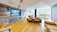 Located at 333 E 11th Ave in Vancouver's Mount Pleasant neighbourhood, this single-floor living space offers a thoughtfully curated lounge environment with a distinct artistic character. The living room is defined by warm hardwood flooring that runs throughout, contributing to a bright and welcoming ambiance.

One of the most striking features is the expansive gallery wall, where an eclectic collection of framed artwork covers nearly every wall surface, creating a visually dynamic and culturally rich atmosphere. A large-format television is mounted on a reclaimed wood-clad accent wall, which also houses a sleek linear fireplace, serving as the focal point of the main seating area.

Floor-to-ceiling glazed windows flood the space with natural light and frame elevated views of the surrounding Vancouver cityscape and greenery, with a bold cylindrical structural column adding an industrial design element. The lounge is furnished with multiple seating arrangements, including a generously sized sofa, accent chairs, and a bistro-style dining nook positioned near the windows, ideal for casual gatherings.

A panelled grey feature wall with refined wainscoting detail adds a touch of classic elegance to one section of the room, complemented by a small integrated beverage station nearby. Recessed lighting and pendant fixtures provide layered illumination, completing this versatile and design-forward space.