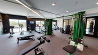 This modern fitness center is thoughtfully designed to support a full spectrum of workout routines. The space features a tray ceiling with recessed lighting and integrated linear LED strips that create a bright, energizing atmosphere, complemented by large windows that invite natural light and outdoor views.

The main workout floor is carpeted and organized into distinct training zones. The cardio area includes multiple treadmills, elliptical machines, and a stationary bike, all positioned near the windows to maximize natural light. At the center of the space stands a substantial multi-station cable machine finished in bold green, serving as the anchor for the strength training zone.

A dedicated free weights area houses an extensive rack of dumbbells, paired with adjustable benches for a variety of exercises. A power rack with an Olympic bench is positioned against a striking diagonal-striped accent wall, ideal for heavy lifting. A leg press machine and additional cable attachments round out the strength equipment selection.

Floor-to-ceiling mirrors line multiple walls, visually expanding the space and allowing users to monitor their form. A motivational wall mural adds an inspiring touch. The fitness center is accessed through sleek glass-panel doors, and a separate enclosed area with glass walls provides additional functional training space.