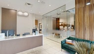 Located at 22555 SW 107th Ave in Goulds, Florida, this single-floor professional office space offers a thoughtfully designed layout comprising two private offices, two utility rooms, a kitchen, and a welcoming entrance. The interior is defined by a clean, contemporary aesthetic with large-format porcelain tile flooring, recessed lighting throughout, and a neutral color palette of whites and warm beiges accented by rich wood paneling.

The entrance greets visitors with a stylish waiting area featuring a teal velvet sofa, leather accent chair, a glass-top coffee table, and a striking sputnik-style chandelier. Floor-to-ceiling windows with roller shades bathe the space in natural light while offering views of the surrounding landscaping. A sleek, fluted-panel reception desk serves as the operational centerpiece, complemented by a bold decorative wall sconce and warm wood accent panels.

The two private offices are enclosed by full-height frameless glass partitions, maintaining an open, airy feel while providing privacy. One office features a large-scale photographic mural, adding a distinctive visual element. The kitchen and utility rooms support day-to-day operations efficiently. Throughout, the space balances professional functionality with polished, modern design details.