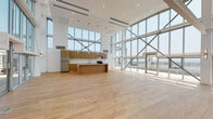 Located at 8155 Van Nuys Blvd in Panorama City, CA, this impressive two-story space delivers a bold architectural statement defined by dramatic floor-to-ceiling curtain glass walls that flood the interior with natural light and frame expansive views of the surrounding San Fernando Valley skyline. The upper level serves as the main living and kitchen area, showcasing soaring double-height ceilings, light hardwood flooring, and a sleek kitchen outfitted with warm wood cabinetry, white quartz countertops, stainless steel appliances including a refrigerator and dishwasher, and a stainless undermount sink. Structural cross-bracing elements integrated into the glass façade add an industrial yet refined aesthetic character. Direct access to an outdoor terrace extends the living experience with open-air views across the valley. A modern staircase with dark metal railings and horizontal balusters connects the two levels with architectural elegance. The lower level features polished concrete floors, crisp white walls with wall-mounted sconce lighting, and versatile open rooms suited for a variety of uses. A clean, tiled bathroom with a soaking tub, shower, and sliding glass enclosure completes the space. Throughout, the design emphasizes openness, natural light, and contemporary finishes.