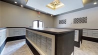 The Mail Center is a thoughtfully designed communal space that balances functionality with polished aesthetics. At its core sits a large, dark-finished central island housing rows of individual resident mailboxes with sleek metallic fronts, providing convenient and organized mail access. Additional mailbox units line the perimeter walls, ensuring ample capacity for the building's residents.

The room's architectural character is defined by a coffered skylight at the ceiling's center, fitted with a distinctive starburst light fixture that serves as a focal point and bathes the space in warm ambient light. Recessed lighting throughout the ceiling further enhances visibility and contributes to the room's bright, welcoming atmosphere.

Neutral gray walls provide a clean backdrop, accented by decorative checkered metal wall art pieces that add a contemporary touch. An arched window allows natural light to filter into the space, complementing the artificial lighting. The light-toned tile flooring ties the design together, creating a cohesive and modern environment that is both practical and visually refined.