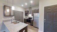 This single-story, 2-bedroom, 2-bathroom residence offers a thoughtfully designed layout with modern finishes throughout. The kitchen serves as a standout feature, showcasing dark gray shaker-style cabinetry paired with light granite countertops that provide a striking contrast. A stainless steel appliance suite includes a French door refrigerator, electric range, and built-in microwave, while a sleek wall-mounted range hood adds both function and a contemporary design element. The kitchen island offers additional counter space and is equipped with a stainless steel undermount sink, making meal prep and cleanup convenient. Light wood-look flooring runs throughout, contributing to a clean, cohesive aesthetic. Recessed lighting and a pendant light above the island ensure the kitchen is well-lit. A large double-door closet near the kitchen provides generous storage. The neutral color palette of white walls and gray tones creates a bright, versatile backdrop throughout the home. The overall layout is efficient and well-organized, making the most of the available space across all rooms.