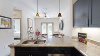 This well-appointed single-floor residence offers a thoughtfully designed open-concept layout that seamlessly connects the kitchen, dining, and living areas. The kitchen is a standout feature, showcasing dark espresso cabinetry, granite countertops, stainless steel appliances including a refrigerator, range, and microwave, and a peninsula with bar seating illuminated by warm pendant lighting. Wood-style flooring runs throughout the main living areas, lending a cohesive and modern aesthetic. The living room is bright and inviting, benefiting from large windows and glass French doors that flood the space with natural light and provide outdoor access. A ceiling fan adds comfort to the open living area. Two bedrooms offer a calm retreat, each carpeted and filled with natural light from generous windows fitted with blinds. The full bathroom is well-equipped with a granite-topped vanity featuring dark cabinetry, wood-style flooring, and a full bathtub with shower. Clean white walls and neutral tones throughout create a bright, airy ambiance that complements the contemporary finishes, resulting in a cohesive and functional living space.