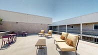 Located at 742 S Westlake Ave in the Westlake neighborhood of Los Angeles, this rooftop terrace offers an expansive open-air retreat with sweeping views of the surrounding cityscape and iconic California sky. The deck is finished in smooth concrete and bathed in abundant Southern California sunlight, creating a bright and inviting atmosphere throughout the day.

The terrace is thoughtfully furnished with multiple distinct seating areas to accommodate a variety of uses. Cushioned lounge chairs and settees with warm neutral upholstery provide comfortable spots for relaxing, while a slatted wood coffee table adds a natural accent. A vibrant teal side table introduces a pop of color to one seating vignette, and a bold purple bistro table with matching chairs creates a lively social corner ideal for casual dining or conversation.

Perimeter railing along one side of the deck offers unobstructed views of the Los Angeles skyline and neighboring palm trees, reinforcing the quintessential L.A. outdoor experience. Stucco-finished accent walls border portions of the terrace, featuring wall-mounted sconce lighting for evening ambiance. Planters with greenery add a touch of nature to the space. The terrace's open layout and versatile furnishings make it well-suited for both quiet relaxation and social gatherings in an urban rooftop setting.