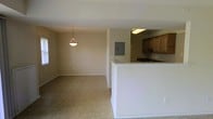 This single-story space features an open-concept design that integrates the kitchen and living area into one cohesive layout. The living area is finished with neutral-toned tile flooring and painted walls in a soft beige palette, contributing to a clean and understated ambiance. A pendant light fixture hangs centrally, providing focused overhead lighting to the space. Natural light filters in through a window along the far wall, brightening the interior.

The kitchen is defined by a white peninsula counter that serves as a natural divider between the cooking and living zones. Medium-toned wooden cabinetry lines the upper kitchen walls, offering ample storage. A dark-toned sink is set into the counter, and under-cabinet lighting provides practical illumination for food preparation. An electrical panel is mounted on the wall adjacent to the kitchen, discretely integrated into the layout.

The overall design is straightforward and functional, with minimal ornamentation. The open flow between the kitchen and living space makes the interior feel connected and accessible, suitable for everyday living.