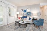 This thoughtfully designed single-floor residence offers a seamless blend of comfort and functionality across its well-defined living spaces. The living room serves as the heart of the home, featuring plush carpeting, a patio door with patterned drapes that invite abundant natural light, and a curated gallery wall displaying an eclectic mix of framed artwork. The open-concept kitchen is equipped with light maple cabinetry, granite countertops, stainless steel appliances including a refrigerator and microwave, a gas range, and a double sink — all accented by tile flooring and a pass-through breakfast bar that connects seamlessly to the living area. The bedroom is a serene retreat, highlighted by dual windows with sheer curtains that frame lush outdoor greenery, soft carpeting, and warm ambient lighting. A versatile den or office space features soft blue accent walls, carpet flooring, and a large window, making it ideal for remote work or a sitting room. Two bathrooms are finished with light maple vanities, tile flooring, large mirrors, and clean white fixtures — one featuring a shower-tub combination. Throughout the residence, a consistent neutral palette and abundant natural light create a warm, inviting ambiance.