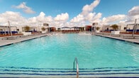 Located at 503 George Bush Dr W in College Station, Texas, this rooftop amenity level offers a well-appointed outdoor destination designed for both relaxation and social gathering. The centerpiece of the space is a generously sized swimming pool with vivid turquoise water, framed by a wide surrounding deck that provides ample room for lounging beneath the open Texas sky.

Adjacent to the pool, a spacious outdoor dining and entertainment area features warm wood-toned deck flooring and numerous seating arrangements with slatted chairs and tables, all sheltered beneath large tensile shade sails in deep navy blue. A dedicated bar area with counter seating is positioned along the perimeter, complemented by built-in grilling stations to support outdoor entertaining. A living green wall displaying the "PW" logo adds a distinctive branded accent to the space.

The rooftop also includes an expansive open-air plaza finished in light aggregate concrete, offering additional gathering space with bench seating and pergola structures. Raised planters with tropical palms and manicured greenery are integrated throughout, lending a lush, resort-like atmosphere. Panoramic views of the surrounding College Station skyline are visible from multiple vantage points, enhancing the elevated outdoor experience this rooftop amenity provides.