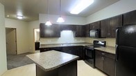 This single-floor space showcases a well-appointed kitchen as its centerpiece. The kitchen is outfitted with rich, dark espresso-toned cabinetry that extends to near the ceiling, providing generous storage capacity. Granite countertops in a light, speckled finish run along the perimeter and are complemented by a matching central island, offering ample prep and workspace. Black appliances, including a range, built-in microwave, and dishwasher, are seamlessly integrated into the cabinetry for a cohesive, contemporary look. A double-basin sink is positioned along the countertop run, conveniently accessible within the kitchen's layout. Two pendant lights hang above the island, providing focused task lighting, while a skylight or flush ceiling fixture brightens the space with additional overhead illumination. The kitchen's layout follows an efficient L-shape with the island adding versatility. Light-toned flooring contrasts with the dark cabinetry, enhancing the brightness of the space. A doorway visible in the background hints at additional rooms, suggesting a connected and functional floor plan throughout the residence.