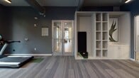 Located at 1020 E Broad St in Savannah, Georgia, this single-floor fitness center blends industrial aesthetics with modern functionality. Exposed brick columns anchor the open floor plan, complemented by dark slate-blue walls, patterned carpet tiles with green accent zones, and visible ductwork overhead that reinforce the space's contemporary industrial character. Large windows allow natural light to filter in, offering views of the surrounding greenery and contributing to an energizing atmosphere.

The cardio zone is outfitted with elliptical trainers, treadmills, and a stationary bike, each equipped with personal display screens. A dedicated strength training area features a squat rack loaded with a barbell, along with a full dumbbell rack displayed in front of floor-to-ceiling mirrors that visually expand the space. A functional training corner houses a boxing/combat training station with red punch targets and interactive display, alongside additional cable and resistance machines.

A built-in open shelving unit near the entrance provides organized storage for equipment such as yoga mats and balance boards. The entry area features glass double doors connecting the fitness center to adjacent amenity spaces. Ceiling fans ensure comfortable airflow throughout, while wall-mounted televisions and linear LED lighting add to the polished, well-appointed finish of this comprehensive fitness facility.