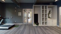 Located at 1020 E Broad St in Savannah, Georgia, this single-floor fitness center blends industrial aesthetics with modern functionality. Exposed brick columns anchor the open floor plan, complemented by dark slate-blue walls, patterned carpet tiles with green accent zones, and visible ductwork overhead that reinforce the space's contemporary industrial character. Large windows allow natural light to filter in, offering views of the surrounding greenery and contributing to an energizing atmosphere.

The cardio zone is outfitted with elliptical trainers, treadmills, and a stationary bike, each equipped with personal display screens. A dedicated strength training area features a squat rack loaded with a barbell, along with a full dumbbell rack displayed in front of floor-to-ceiling mirrors that visually expand the space. A functional training corner houses a boxing/combat training station with red punch targets and interactive display, alongside additional cable and resistance machines.

A built-in open shelving unit near the entrance provides organized storage for equipment such as yoga mats and balance boards. The entry area features glass double doors connecting the fitness center to adjacent amenity spaces. Ceiling fans ensure comfortable airflow throughout, while wall-mounted televisions and linear LED lighting add to the polished, well-appointed finish of this comprehensive fitness facility.