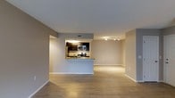 This single-floor space features an open-concept design that seamlessly connects the kitchen, dining, and living areas into one cohesive layout. The kitchen is positioned at the heart of the unit, framed by dark cabinetry that contrasts sharply against light-toned walls. A granite-topped peninsula serves as both a functional workspace and a natural divider between the kitchen and the main living area, offering casual seating potential and added counter space. Stainless steel appliances, including a range, microwave, and refrigerator, are built into the kitchen setup, complemented by a tile backsplash that adds texture. Track lighting illuminates the living area, while recessed lighting within the kitchen ensures consistent brightness throughout. Light hardwood-style flooring runs continuously across the space, contributing to a clean, unified aesthetic. The neutral warm-gray wall palette enhances the open feel and provides a versatile backdrop. HVAC vents are integrated into the ceiling, suggesting a climate-controlled environment. The overall ambiance is modern and functional, with an efficient use of space throughout.