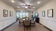 This thoughtfully designed single-floor communal space offers a welcoming and functional environment across several distinct areas. The main gathering room is a bright, open space featuring ceiling fans, recessed lighting, and large windows that flood the room with natural light. It is furnished with round tables and upholstered chairs arranged to encourage social interaction, alongside a large flat-screen television mounted on a dark wood media console. Decorative wainscoting lines the lower walls throughout, lending a refined, classic character to the interiors.

Adjacent to the main room is a more intimate game and media nook, where a round dark-wood table is set with a board game and surrounded by cushioned chairs. Large windows bring in ample daylight, while a flat-screen TV and storage cabinet complete the space, making it ideal for leisure activities.

The corridors are wide and carpeted, with consistent wainscoting and framed artwork creating a polished, gallery-like feel. Seating alcoves are thoughtfully placed along the hallway, furnished with patterned armchairs, small side tables with greenery, and decorative mirrors, offering quiet spots for relaxation. An elevator landing serves as a functional hub connecting the space. Overall, the design balances comfort and elegance, creating an inviting atmosphere throughout.