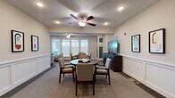 This thoughtfully designed single-floor communal space offers a welcoming and functional environment across several distinct areas. The main gathering room is a bright, open space featuring ceiling fans, recessed lighting, and large windows that flood the room with natural light. It is furnished with round tables and upholstered chairs arranged to encourage social interaction, alongside a large flat-screen television mounted on a dark wood media console. Decorative wainscoting lines the lower walls throughout, lending a refined, classic character to the interiors.

Adjacent to the main room is a more intimate game and media nook, where a round dark-wood table is set with a board game and surrounded by cushioned chairs. Large windows bring in ample daylight, while a flat-screen TV and storage cabinet complete the space, making it ideal for leisure activities.

The corridors are wide and carpeted, with consistent wainscoting and framed artwork creating a polished, gallery-like feel. Seating alcoves are thoughtfully placed along the hallway, furnished with patterned armchairs, small side tables with greenery, and decorative mirrors, offering quiet spots for relaxation. An elevator landing serves as a functional hub connecting the space. Overall, the design balances comfort and elegance, creating an inviting atmosphere throughout.