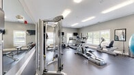 Located at Sportsman Park Lane in Fort Wayne, Indiana, this fitness center offers a dedicated exercise room designed to support a well-rounded workout routine. The space features durable dark flooring throughout, complemented by neutral gray walls and bright recessed fluorescent lighting that keeps the room well-illuminated at all times. A large floor-to-ceiling mirror spans one wall, visually expanding the space and providing users with proper form feedback during exercise. Natural light flows in through multiple windows along the exterior walls, offering pleasant outdoor views of the surrounding grounds. The equipment selection is comprehensive, including treadmills, an elliptical trainer, a stair climber, and a rowing machine for cardiovascular workouts. Strength training is supported by a cable machine, a weight bench, and a full rack of dumbbells. Stability balls and additional accessories are neatly organized on a dedicated rack. Multiple wall-mounted flat-screen televisions are positioned throughout the room, ensuring entertainment is accessible from various workout stations. The overall layout is efficient and thoughtfully arranged, allowing for comfortable movement between equipment. The clean, modern aesthetic and practical amenities make this fitness center a functional and inviting space for daily exercise.