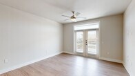 This well-appointed single-floor residence offers a clean, modern aesthetic throughout, characterized by a neutral color palette, light wood-style flooring, and abundant natural light. The open-concept kitchen serves as a central hub, featuring white shaker cabinetry, a subway tile backsplash, stainless steel appliances including a refrigerator, microwave, and range, and a large island with a light quartz countertop illuminated by a trio of pendant lights. Recessed lighting and a connected entry area complete this functional space. The main living area is spacious and airy, highlighted by French doors fitted with transom windows above, allowing generous natural light and outdoor views. A ceiling fan with a brushed nickel finish adds comfort overhead. Additional rooms feature plush carpeted flooring, ceiling fans, and large windows with horizontal blinds that offer views of the surrounding neighborhood. The bathroom is well-designed with a white vanity offering ample cabinet storage, gold-toned hardware, a large mirror, and wood-style flooring. Conveniently adjacent is a walk-in closet with built-in shelving and hanging rods, providing practical storage solutions. The overall ambiance is clean, bright, and move-in ready.