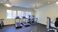 This fitness center offers a focused and functional workout environment thoughtfully designed to meet everyday exercise needs. The space is outfitted with multiple treadmills and elliptical cross-trainers, providing a solid selection of cardio equipment for users of varying fitness levels. Additional exercise equipment, including a weight bench, is also present, allowing for a well-rounded workout routine.

The room features a durable dark rubberized flooring, ideal for high-traffic fitness use, while the clean white walls lend a bright and open feel to the interior. A decorative pendant light fixture hangs from the ceiling, complementing the recessed lighting above to ensure the space is well-lit at all times. Large windows along one wall allow ample natural light to fill the room, offering views of the surrounding building exterior and keeping the atmosphere fresh and inviting.

The single-floor layout is straightforward and accessible, maximizing the usable floor area for equipment placement and free movement. Overall, this fitness center strikes a balance between practicality and comfort, providing residents or users with a convenient, on-site space to maintain an active and healthy lifestyle.