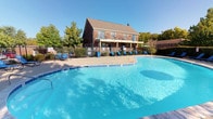 This outdoor amenity area centers around a large, freeform swimming pool with clear blue water, designed to accommodate residents seeking recreation and relaxation. The pool's curvilinear shape adds visual interest and provides ample room for swimming. Surrounding the pool is a broad concrete deck finished with tile edging, furnished with multiple blue lounge chairs that offer comfortable spots for sunbathing and leisure throughout the day.

A black wrought iron fence encloses the pool area, providing both security and a defined boundary while maintaining an open, airy feel. A shaded umbrella seating area is positioned along one side, offering a retreat from direct sun exposure.

The backdrop is anchored by a two-story brick building with a gabled roof, covered lower-level porch, and multiple windows that overlook the pool area. Mature trees and green landscaping frame the perimeter, contributing a natural, park-like atmosphere. The combination of classic brick architecture, well-maintained grounds, and thoughtfully arranged outdoor furnishings creates an inviting communal environment ideal for warm-weather enjoyment.