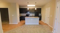 This single-story space showcases an open-concept kitchen that serves as the focal point of the layout. The kitchen is outfitted with dark espresso-toned cabinetry that extends to the upper and lower walls, providing generous storage capacity. Granite countertops in warm beige and gold tones offer a striking contrast against the dark cabinets and are carried onto the central kitchen island, which also features a built-in sink and an integrated electrical outlet for added functionality.

Two pendant lights hang above the island, providing focused task lighting, while a flush-mount fixture overhead ensures the space remains well-lit. Black appliances, including a range, microwave, and refrigerator, complement the dark cabinetry for a cohesive, contemporary aesthetic.

The flooring transitions from hard surface tile in the kitchen to plush neutral carpeting in the adjoining living area, visually defining each zone while maintaining an open, connected feel. Light green-toned walls contribute a soft, neutral backdrop throughout. Multiple doorways visible from the main space suggest access to additional rooms, reinforcing the functional and practical layout of this residence.