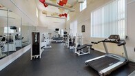 Located at 4815 Garden Ranch Dr in Colorado Springs, Colorado, this single-floor fitness center is a comprehensively equipped facility designed to support a full range of workout routines. The main gym area is characterized by soaring ceilings with clerestory windows that flood the space with natural light, creating an open and energizing atmosphere. Decorative red and neutral fabric elements suspended from the ceiling add a distinctive visual character to the room. The floor is covered in durable dark rubber matting, ideal for heavy equipment and high-impact activities. The strength training section features a variety of cable machines, free weights, dumbbells, and weight benches, while the cardio area includes treadmills, elliptical trainers, and stationary bikes. Full-length mirrors along one wall enhance the sense of space and allow for proper form monitoring. A secondary area, finished with light tile flooring, serves as a transitional lounge and cardio zone, complete with mounted televisions, a beverage vending station, and small café-style tables and chairs. Large floor-to-ceiling windows in this area provide views of an outdoor patio space. A water fountain station and access to locker room facilities are also available, rounding out the amenities of this well-appointed fitness center.