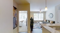 This single-floor residence offers a thoughtfully arranged layout that seamlessly connects its primary living spaces. The open-plan living area is bright and welcoming, benefiting from large windows dressed with white blinds that allow generous natural light to fill the room. A ceiling fan overhead provides comfort and airflow, while hardwood-style flooring anchors the space with a clean, contemporary aesthetic. The living area is complemented by a geometric-patterned area rug, a glass coffee table, and a comfortable sofa, creating an inviting atmosphere for relaxation.

Adjacent to the living area, a kitchen counter with granite-style surfaces and a pendant light fixture hints at a functional kitchen space nearby. A bold, large-scale artwork displayed in the hallway adds a striking artistic touch to the interior.

The bedroom, visible through a doorway, is carpeted for warmth and comfort, and features its own ceiling fan and windows that offer views of lush greenery outside. The neutral, warm wall tones throughout the unit create a cohesive and calming ambiance, making the space feel polished and livable.