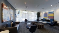 This single-floor space presents a thoughtfully designed lounge area that blends modern aesthetics with functional comfort. Floor-to-ceiling sheer white curtains line an expansive window wall, flooding the interior with soft, diffused natural light while maintaining a sense of openness and connection to the outdoors, where mature trees are visible beyond the glass.

The seating arrangement is diverse and carefully curated, featuring sleek low-profile sofas, ergonomic swivel chairs, and patterned ottomans in a bold checkered design, creating visual interest across the space. A cluster of small round accent tables provides practical surface areas throughout.

A striking pendant light installation anchors one corner of the room, showcasing a collection of teardrop-shaped blue and clear glass pendants suspended at varying heights, adding an artistic focal point. Recessed ceiling lighting complements this decorative feature, ensuring the space remains well-lit throughout.

A tall indoor potted tree introduces a natural element near the window wall, softening the contemporary environment. The carpeted flooring in a textured charcoal pattern ties together the neutral color palette, while a colorful wall-mounted art panel adds a subtle pop of color to one end of the room.