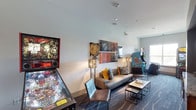 This single-level arcade is designed to deliver an engaging, entertainment-focused environment that blends retro gaming culture with modern lounge aesthetics. The space is laid out along a elongated open floor plan, with patterned carpet in deep blue and grey tones running throughout, adding visual texture underfoot.

At one end, classic stand-up arcade cabinets and a pinball machine anchor the gaming experience, providing a nostalgic focal point. The lounge area runs alongside, featuring a grey upholstered sofa accented with bold mustard and patterned throw pillows, complemented by leather sling chairs and modular nesting coffee tables. Tall metallic floor lamps provide warm ambient lighting, while a curated gallery-style wall arrangement of mixed-media artwork adds an artistic edge.

At the opposite end, a built-in media nook takes center stage, showcasing a large flat-screen TV mounted within a bold mustard-yellow recessed wall. Below it, wood-toned cabinetry with granite countertops and an under-counter beverage refrigerator provides a functional and stylish serving area. Natural light enters through windows at the far end, brightening the space. Recessed ceiling lighting throughout ensures the entire room is well-illuminated.